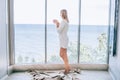 Slim woman in white knitted home dress enjoying sea view in hotel room next to big window with a cup of coffee. Vacation Royalty Free Stock Photo