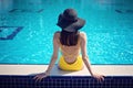 Slim woman in yellow swimsuit and hat sitting on the edge of the pool playing with water feet, back view Royalty Free Stock Photo