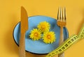 Slimming concept: yellow dandelion flowers lie on a blue plate, next to a fork, centimeter, top view, yellow background, space for Royalty Free Stock Photo