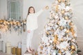 Small adorable female child in white sweater and trousers holds toy for decoration, decorates New Year tree. Cheerful little kid n