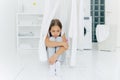 Small adorable girl sits on floor, being punished by parents, poses near clothes dryer, focused down with sad expression, washing Royalty Free Stock Photo