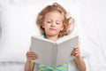 Small appealing female kid rests in bed with book, reads interesting exciting story before sleep, looks attentively at pages, lies Royalty Free Stock Photo