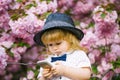 Small baby boy in white shirt and retro hat with bow tie with blonde hair playing on mobile phone in spring pink flower Royalty Free Stock Photo