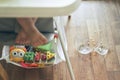 A small baby on a high chair dropped a cup of noodle soup on the wooden floor. The transparent plate is divided into two