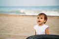 Small baby, little girl in white t-shirt sitting in sand at the beach near to the old tire of a vehicle. Ecological problems