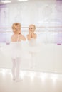 Small ballerina girl in white tutu standing next to the mirror with her reflection in it at dance studio. Children and ballet