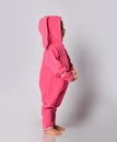 Positive barefoot cute blonde baby girl in pink warm comfortable jumpsuit with hood and bunny ear stands side to camera
