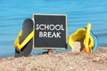 Small black chalkboard with text School Break, beach toys and flip flops on sand near sea. Seasonal holidays