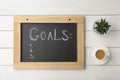 Small blackboard with empty list of goals, houseplant and cup of coffee on white wooden table, flat lay. Space for text