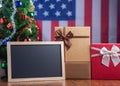 Small blackboard and gift boxes on wooden table with a Christmas tree and American flag in the background. Space for text. Concept Royalty Free Stock Photo