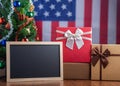 Small blackboard and gift boxes on wooden table with a Christmas tree and American flag in the background. Space for text. Concept Royalty Free Stock Photo