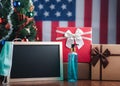 Small blackboard, mask, alcohol gel, and gift boxes on wooden table with a Christmas tree and American flag in the background. Royalty Free Stock Photo