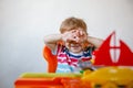 A small blond boy at home sits at an children's table among plastic toys and hides covering his face with his hands Royalty Free Stock Photo