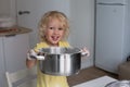 Small blond curly girl in the kitchen holding big metal pot with daugh. Girl is happily smiling, flour is on her hands