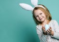 Small blond happy girl in grey cosy home clothing and decorative fur ears standing and holding pastel blue egg