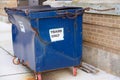 Small blue dented dumpster on wheels on a sidewalk chained to a brown brick wall