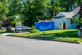 A small blue dumpster a the driveway in front of a house. Two refrigerators are in front of the dumpster