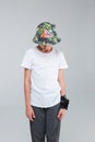 A small boy, in gray sports pants, a white T-shirt and a colored hat, stands with head down. Gray background. Royalty Free Stock Photo