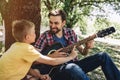 Small boy is leeaning to father`s guitar. He wants to play on it. Bearded guy is holding this guitar and smiling to son