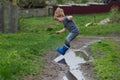 a small boy in rubber boots jumps through a muddy puddle