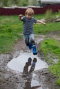 a small boy in rubber boots jumps through a muddy puddle