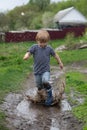 a small boy in rubber boots jumps through a muddy puddle