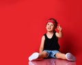 Small boy in stylish casual clothing, hair bandana and white sneakers sitting on floor and feeling excited with hands raised up