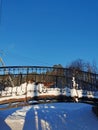 A small bridge with forged cast iron trellises over a frozen river on a sunny winter day against the backdrop of pine trees and a Royalty Free Stock Photo