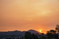 Small bright sun in light orange sunset sky shining behind mountains above houses and green trees Royalty Free Stock Photo