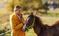 Small brown Arabian horse foal, young woman in yellow orange jacket standing next, blurred sun lit autumn trees background Royalty Free Stock Photo