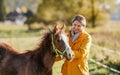 Small brown Arabian horse foal, young woman in yellow orange jacket standing next, blurred sun lit autumn trees background Royalty Free Stock Photo