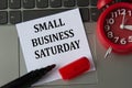 SMALL BUSINESS SATURDAY on a white sheet against the background of the computer keyboard and red clock Royalty Free Stock Photo