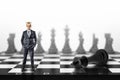 A small businessman with a golden crown on his head stands on a chessboard near a fallen black king.