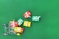 Small cart with scattered gifts on a green background. Red, green and yellow gifts with white bows in a shopping trolley. Royalty Free Stock Photo