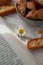 A small chamomile flower lies on the table with a book and cantucci dessert
