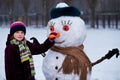 A small cheerful girl holds a big carrot, the nose of a big snowman Royalty Free Stock Photo