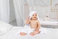 A small child a boy 8 months old is sitting in a towel on a white bed in a light nursery in diapers after bathing Royalty Free Stock Photo