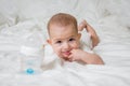 A small child is lying on a white bed on his stomach. The girl put two fingers into her mouth. Nearby there is a plastic bottle Royalty Free Stock Photo