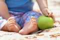 A small child sits on a bedspread with a toy. Bare feet of the baby. Close up of a baby`s heel. child holds a green apple in his h