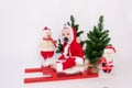 A small child is sitting on a red sleigh on a white isolated background in a Santa costume among Christmas trees, the concept of a Royalty Free Stock Photo