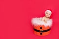 A small child of six months in a Santa hat sits in a basket on a red isolated photophone, a place for text, the concept of the new