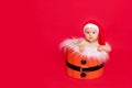 A small child of six months in a Santa hat sits in a basket on a red isolated photophone, a place for text, the concept of the new