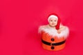 A small child of six months in a Santa hat sits in a basket on a red isolated photophone, a place for text, the concept of the new