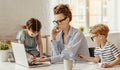 Small children being bored beside tired adult woman working on laptop at home