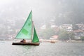 small colorful sailboat with people sitting on it passing row boats and pedal boats on a foggy day with buildings in the