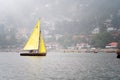 small colorful sailboat with people sitting on it passing row boats and pedal boats on a foggy day with buildings in the