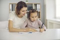 Small concentraited attentive girl sitting with mother or nanny together in room and drawing Royalty Free Stock Photo