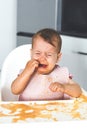 small cute baby 9 months old sits in the kitchen in a high chair and cries, grimy and dirty, spilled food, vertical photo Royalty Free Stock Photo