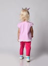 Small cute blonde baby girl kid in pink stylish casual clothing and crown stands back to camera, leaving out