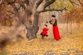 Beautiful mother in a long dress is swinging on a hinged swing little daughter in a red coat in an autumn garden near an old gnarl Royalty Free Stock Photo